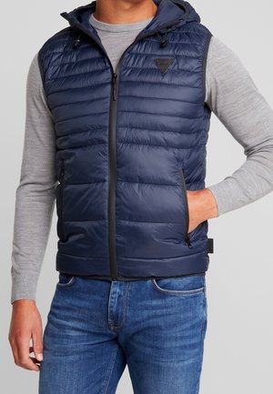 Mand iført en marineblå quiltet vest uden ærmer med hætte over en grå langærmet trøje med hånd i vestlommen, blå jeans synlige.
