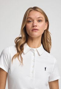 Biała koszulka polo wykonana z bawełny, z kołnierzykiem i pięcioma guzikami. Ma małe haftowane logo na lewej piersi.