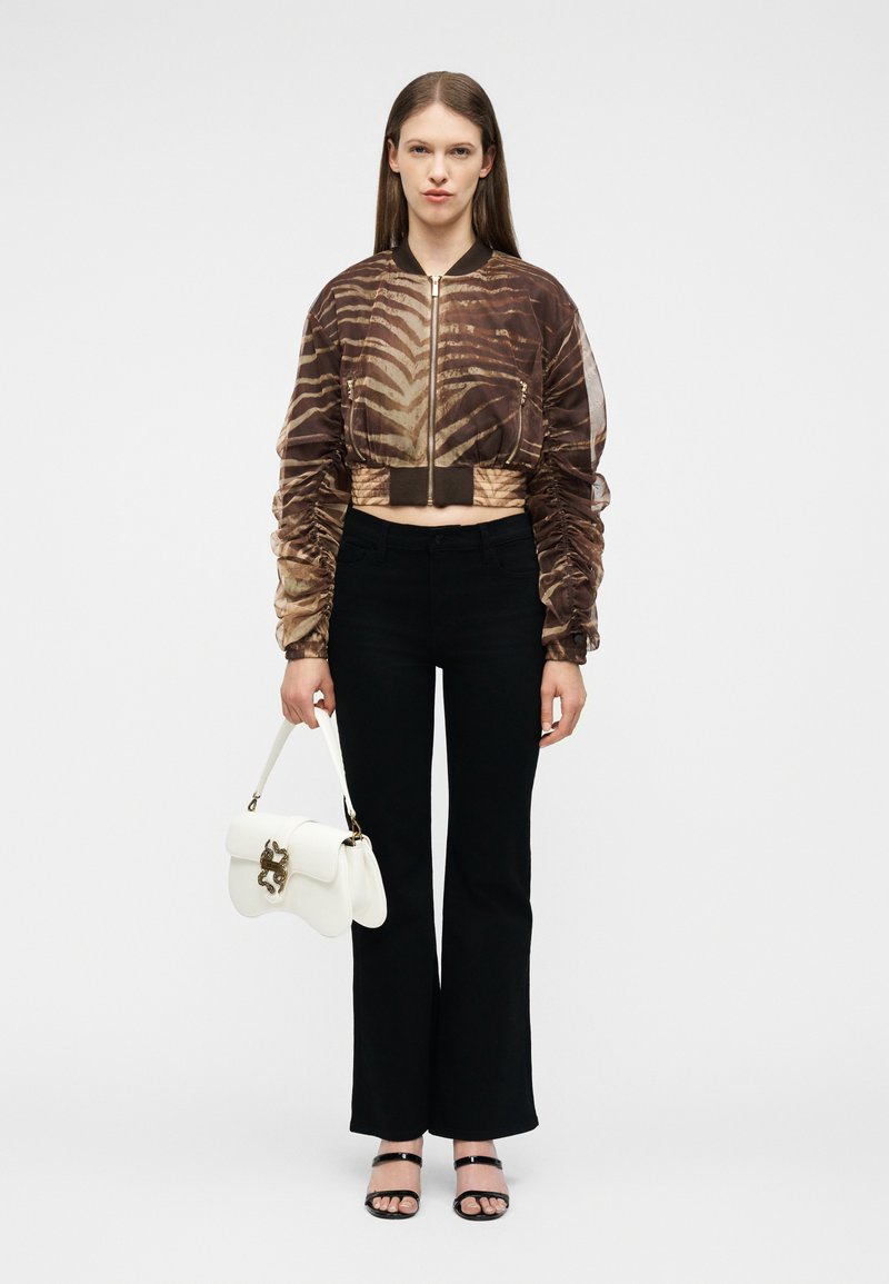 Γυναίκα που στέκεται, φοράει ριγέ καφέ cropped σακάκι, μαύρο παντελόνι, μαύρα σανδάλια και κρατάει μία λευκή τσάντα με χρυσές λεπτομέρειες.