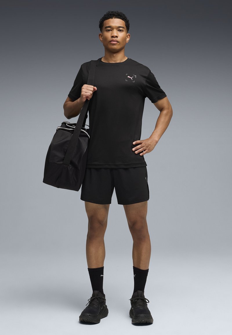 Ung mand står med en sort sportstaske over skulderen, iført sort atletisk t-shirt, shorts, sokker og sneakers på ensfarvet grå baggrund.