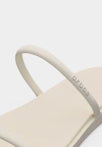 Μπεζ σανδάλι Crocs με λεπτό λουράκι που φέρει ανάγλυφο λογότυπο Crocs σε απλό λευκό φόντο.