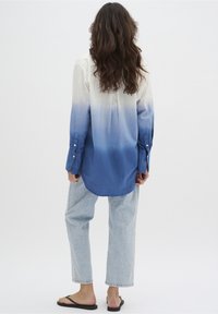 Kvinna med långt vågigt hår står vänd bortåt, klädd i en långärmad skjorta med vit-blå ombre, ljusblå jeans och svarta flip-flop.