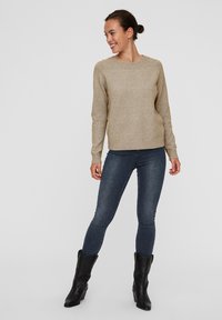 Beige striktrøje med rund halsudskæring og ribbede manchetter, iført tætsiddende blå jeans og sorte ankelstøvler, stående foran en ensfarvet baggrund.