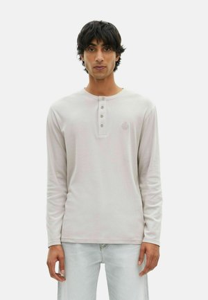 Junger Mann mit dunklem Haar, der ein hellbeiges Langarm-Henley-Shirt und hellen Jeans trägt und vor einem schlichten weißen Hintergrund steht.