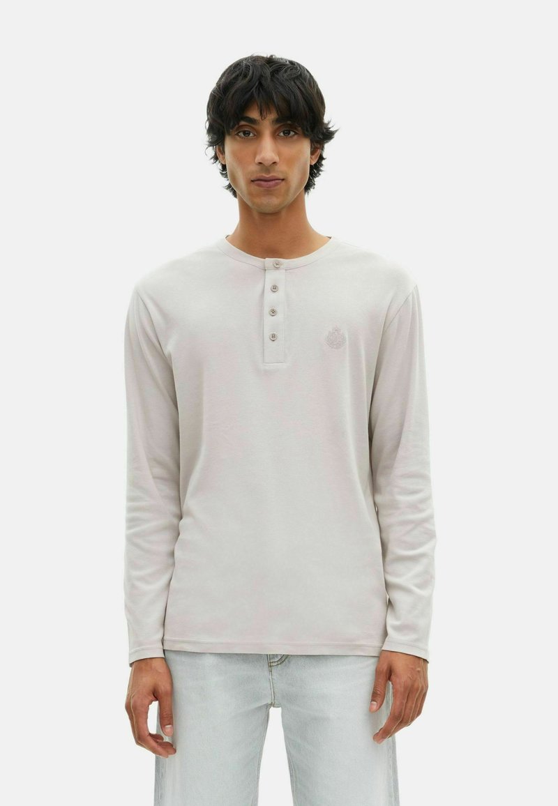Junger Mann mit dunklem Haar, der ein hellbeiges Langarm-Henley-Shirt und hellen Jeans trägt und vor einem schlichten weißen Hintergrund steht.