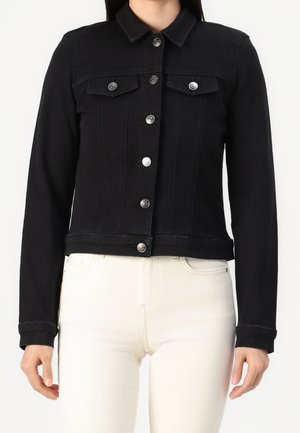 Zwarte spijkerjas met zilveren knopen, borstzakken met klep aan de voorkant, gedragen over witte broek, bovenlichaam en armen zichtbaar.