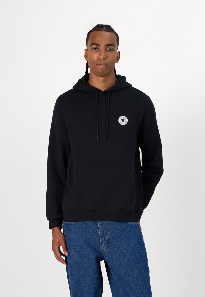 Sudadera negra con capucha, bolsillo frontal y parche de logo circular en el pecho. Combinada con jeans de mezclilla azules.