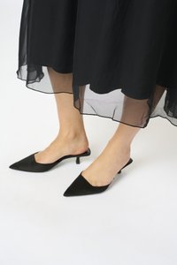 Mule-uri din satin negru cu vârfuri ascuțite și un toc mic, asociate cu o fustă transparentă, cu straturi negre, care are o textură moale și fluidă.