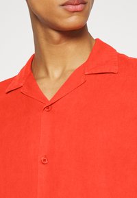 Rode korte mouwen shirt gemaakt van lichte stof, met een platte kraag en twee knopen. De textuur lijkt glad met een subtiele glans.