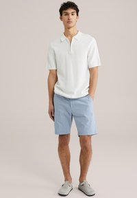 Weißes Polo-Shirt mit kurzen Ärmeln und halbem Reißverschluss, kombiniert mit hellblauen gestreiften Shorts und grauen Slippers vor einem neutralen Hintergrund.