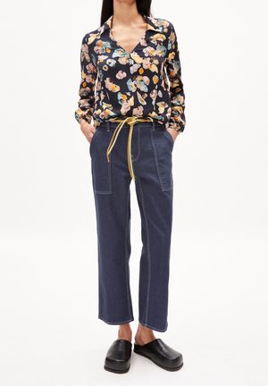 Vrouw draagt een bloemenblouse met lange mouwen, donkerblauwe hoge taille broek met gele riem en zwarte instapschoenen, handen in de zakken.