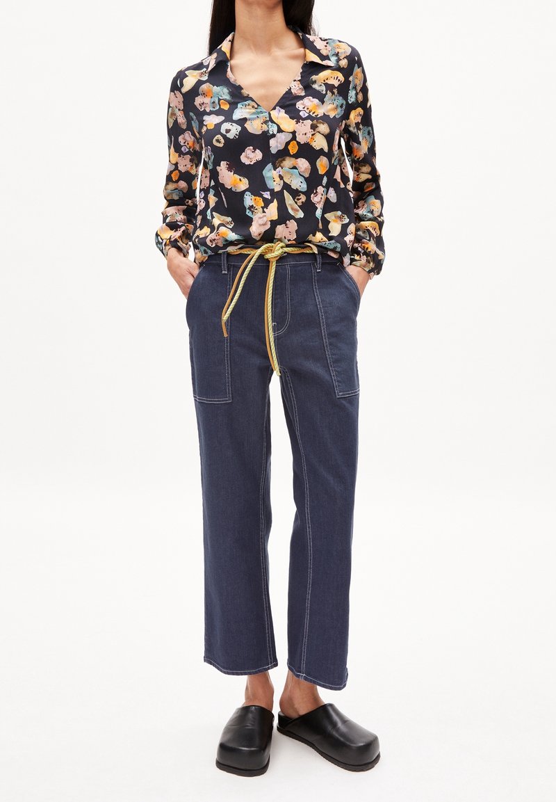 Femme portant un chemisier à manches longues fleuri, un pantalon taille haute bleu foncé avec ceinture jaune et des chaussures noires faciles à enfiler, mains dans les poches.