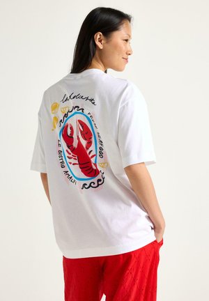 Kvinde iført en hvid T-shirt med en farverig rød hummertil grafik og skaldyrstekst på ryggen, kombineret med røde texturerede bukser.