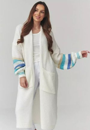 Kobieta ubrana w długi biały dziergany kardigan z dużymi kieszeniami oraz niebieskimi, beżowymi i turkusowymi paskami na rękawach, na białej bluzce i spodniach.