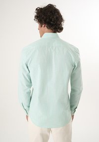 Camisa de cuadros en verde claro, de manga larga, con cuello abotonado y dobladillo curvado, fabricada en una tela suave. Patrón de cuadros visible.