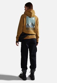 Sudadera amarilla con gráfico de montaña en la parte posterior, combinada con pantalones cargo oscuros y botas negras. Presenta una capucha y puños acanalados.