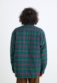 Zöld kockás flanel ing, amely gombos gallérral rendelkezik, lila és sárga részletekkel. Hosszú ujjú és ívelt szegélyű. Puha tapintású.