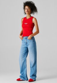 Žena s kudrnatými vlasy, oblečená v červeném tílku Red Bull Racing, světle modré široké džíny a červené boty, stojící s rukama v kapsách.