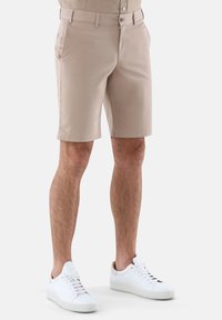 Beige Shorts aus einem glatten Material, mit flacher Vorderseite, Seitentaschen und gesäumten Kanten. Kombiniert mit weißen Sneakers.