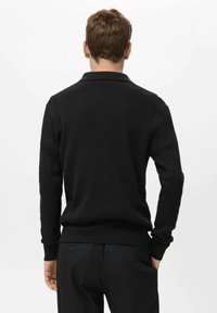 Suéter negro con un diseño de tejido texturizado, mangas largas y puños y dobladillo de canalé. Llevado sobre pantalones oscuros. Vista trasera.