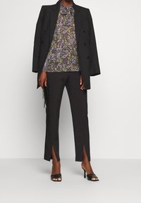 Schwarzer maßgeschneiderter Blazer, gemusterte lila-grün florale Bluse, schmal geschnittene schwarze Hose mit Seitenschlitzen und schwarze offene Sandalen mit Absatz.