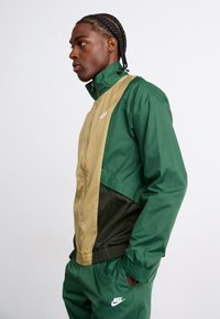 Nike vetrovka v zeleni in peščeni barvi z oprijetim krojem, zadrgo in elastičnimi zapestji. Ima logotip na prsih in kontrastne plošče.