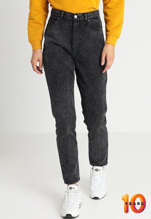 Henkilö pukeutunut korkeavyötäröisiin mustiin happopesuteksareihin, keltaiseen pitkähihainen paitaan ja valkoisiin Nike Air Max -lenkkareihin, seisten yksinkertaisella valkoisella lattialla.