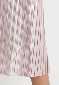 Faltenrock in Hellrosa mit metallischem Glanz. Die strukturierte Oberfläche zeigt vertikale Streifen und einen Saum, der knapp über dem Knie endet.