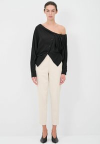 Maglietta nera off-the-shoulder con dettagli a bottoni, abbinata a pantaloni beige a vita alta. Design semplice con vestibilità sartoriale e maniche lunghe.