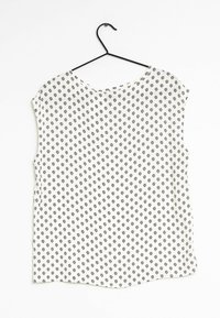 Blusa senza maniche in tessuto bianco con piccolo motivo floreale nero, scollatura tonda e orlo leggermente curvato. Appesa a un gancio nero.