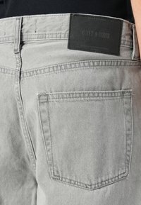 Lys grå denimjeans baklomme med svart merkeetikett "ONLY & SONS" båret av en person mot en svart topp bakgrunn.