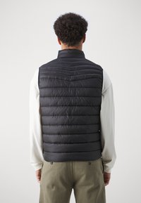 Svart pufferväst med horisontellt quiltat mönster, hög krage och slät textur. Bärs över en ljusfärgad långärmad tröja.