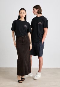 Tricou negru din bumbac cu un grafic colorat "JEANS", asociat cu o fustă lungă din denim închis. Modelul poartă sandale; companionul masculin este îmbrăcat în pantaloni scurți.