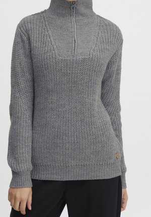 Grauer grob gestrickter Pullover mit hohem Halb-Reißverschluss-Kragen und gerippten Bündchen, getragen mit schwarzen Hosen, Torso und Hände sichtbar.