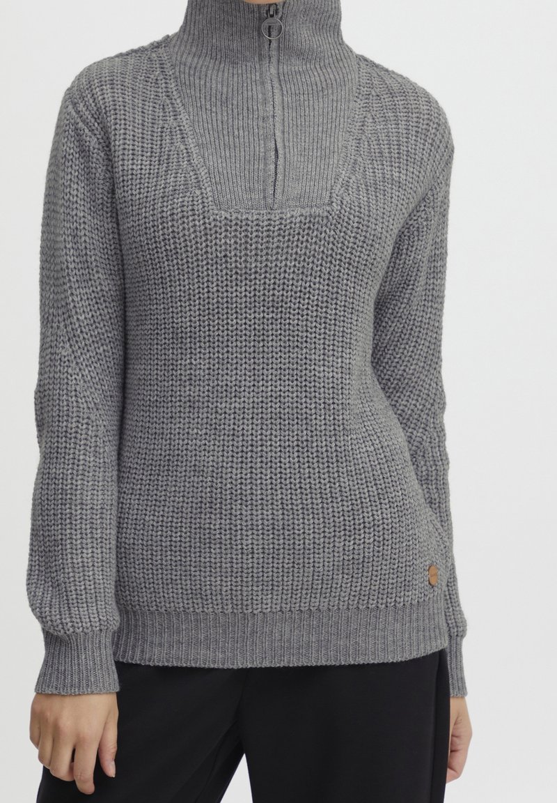 Pull en maille épaisse grise avec col montant à fermeture éclair partielle et poignets côtelés, porté avec un pantalon noir, torse et mains visibles.