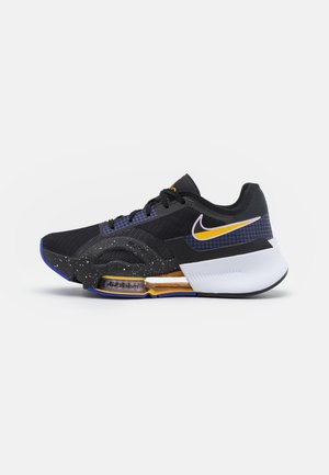 Czarno-biały but do biegania marki Nike z żółtymi akcentami, widoczną podeszwą z poduszką powietrzną, niebieskimi detalami w formie kratki oraz nakrapianą środkową podeszwą na prostym tle.