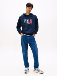 Felpa con cappuccio blu navy con logo bianco e rosso, abbinata a jeans denim larghi blu e sneakers bianche con dettagli blu.