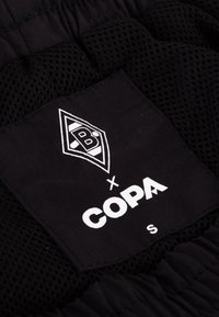Zwarte mesh kledinglabel met het "B x COPA" logo en maat "S" in witte tekst, met een kenmerkende vorm en textuur.