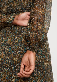 Robe en tissu translucide brun et bleu à motifs, avec des manches amples, des poignets froncés et une finition texturée, mettant en scène une main sur la taille.