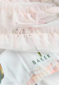 Helltürkiser Tüllstoff mit Rüschenkanten, geschichtet über einem weißen Material mit Blumenmustern und dem Wort "BAKER" in Grün gedruckt.