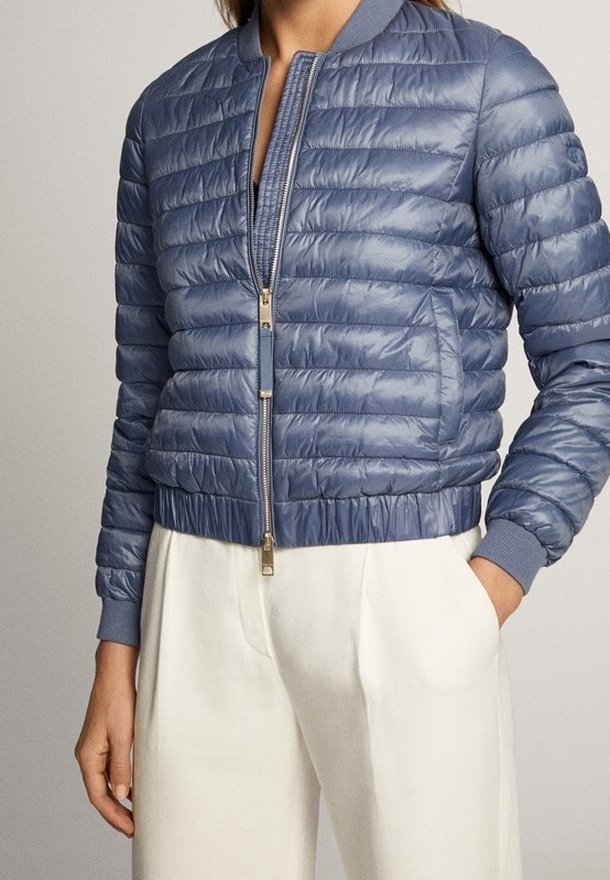 Personne portant un blouson bomber matelassé bleu clair avec une fermeture éclair et un pantalon taille haute ivoire, main dans la poche, debout devant un fond uni.