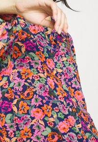 Bloemenblouse in marineblauw met levendige roze, oranje en groene bloemen. Zachte stof, V-hals ontwerp, losse pasvorm. Kenmerkend door knoopdetails.