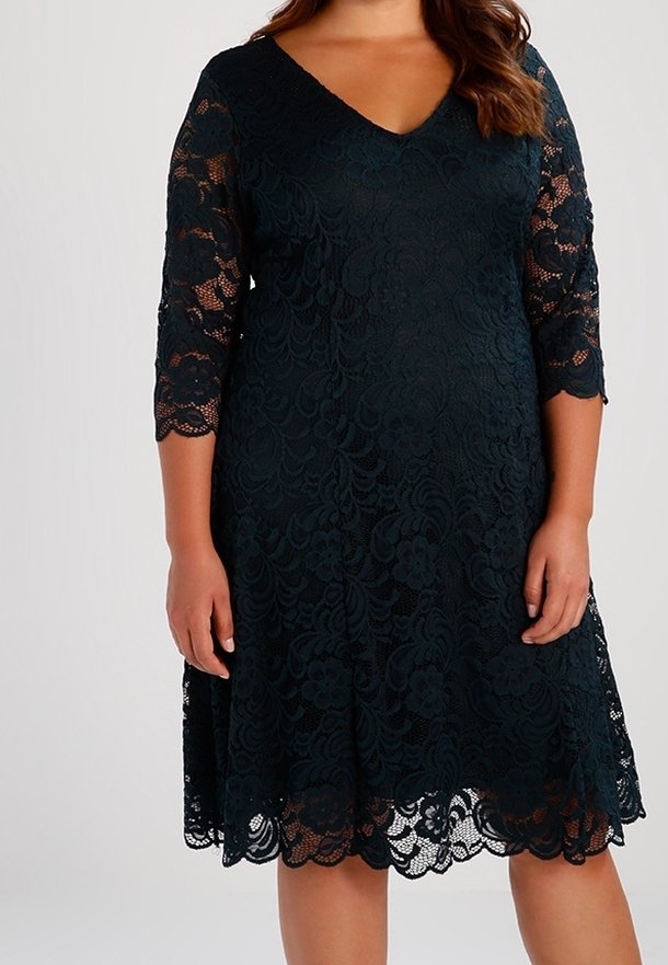 Femme portant une robe en dentelle noire longueur genou, avec des manches trois-quarts et un décolleté en V, debout devant un fond uni.