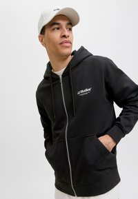 Schwarze Zip-Hoodie mit Kängurutasche, Kordelzügen und weißem Logo auf der Brust. Kombiniert mit einer weißen Mütze, vor einem schlichten Hintergrund.
