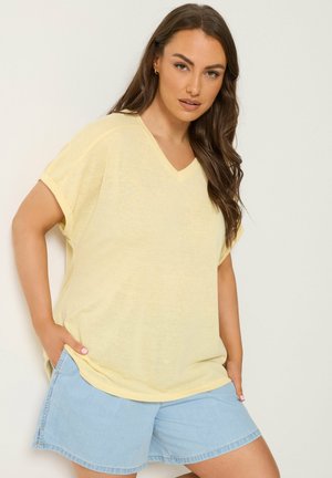 Kvinde med langt brunt hår iført en løs lysegul V-hals T-shirt og lyseblå denimshorts, stående med hænderne i lommerne.