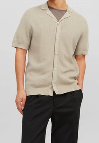 Cardigan a maglia beige a maniche corte con un motivo testurizzato, chiusura con bottoni e colletto piatto. Abbinato a pantaloni neri di taglio ampio.