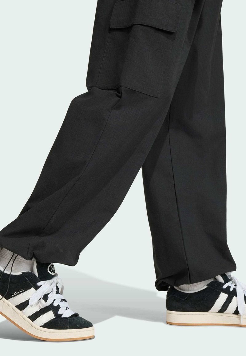 Pantalon cargo noir porté avec des baskets Adidas Campus noires et blanches et des chaussettes blanches, jambes croisées en plein pas sur fond clair.