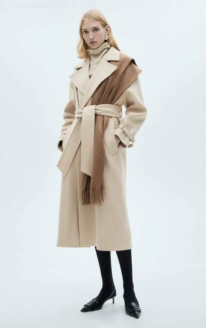 Beige wool coat with a shawl collar, self-tie belt, and brown accent scarf. Model wears black tights and pointed-toe shoes. Smooth texture.