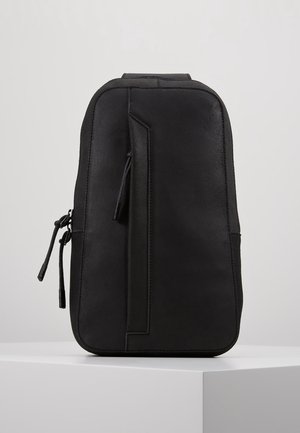 Černá obdélníková kožená kabelka přes rameno s vertikálním zipem a dvěma bočními zipy, stojící na bílém povrchu.