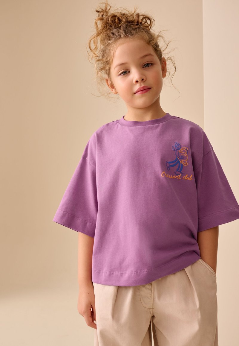 Jeune enfant aux cheveux bouclés portant un t-shirt violet ample avec un petit graphisme et un pantalon beige, debout devant un fond uni.